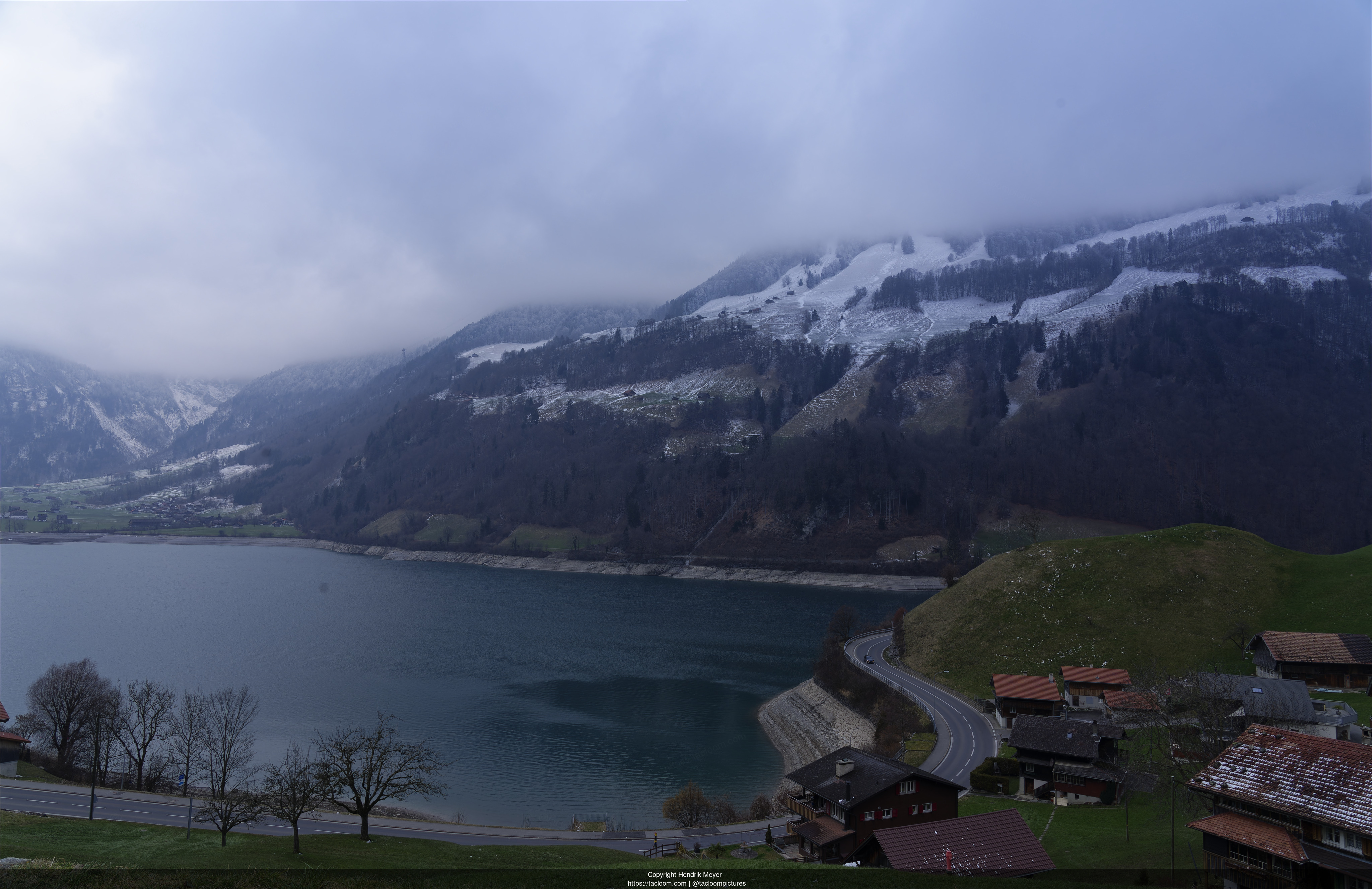 View on a the lake on Lungern. A street weaves from the right to the bottom left of the image. There is a mountain in the background.