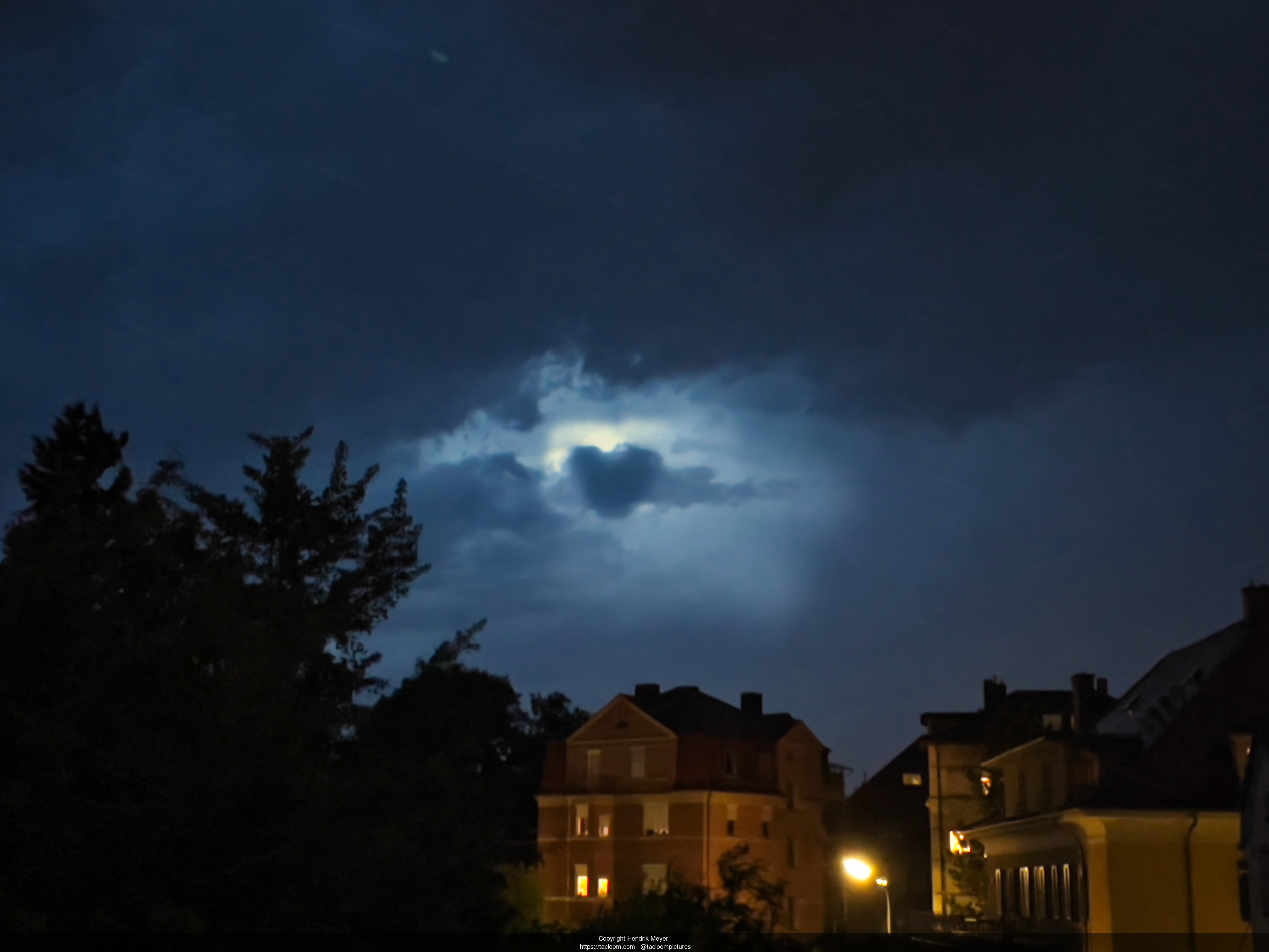 Grim sky, with a heart-shaped cloud in the center. Below the sky there are trees on the left and houses right and center.